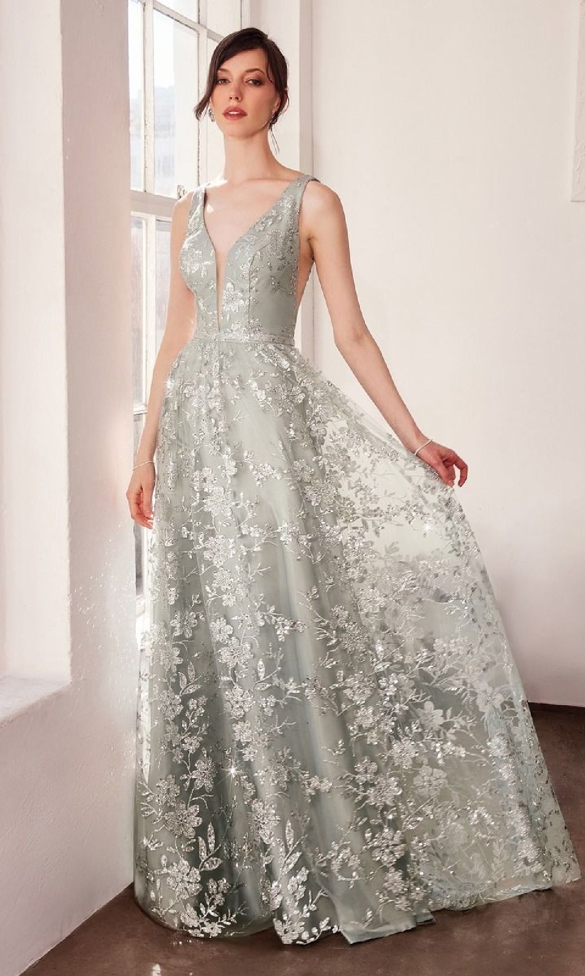 A woman stands by a window wearing the Embroidered-Lace Long Prom Dress: Amoris T606—a sleeveless, floor-length silver gown with a deep V-neck and elegant floral lace detailing.